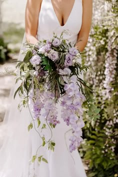 a woman in a wedding dress holding a bouquet of purple flowers and greenery on her shoulder