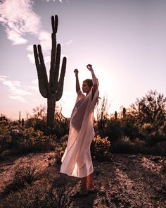 a woman standing in front of a cactus and holding her arms up to the sky