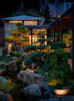 a garden with rocks and trees in front of a house at night, lit up by lights on the windows