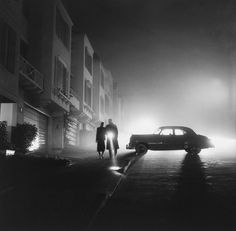 two people are walking down the street at night in front of an old fashioned car