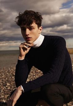 a young man sitting on the beach with his hand under his chin and looking at the camera