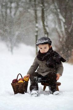 a little boy sitting in the snow with a basket of fruit next to him and looking at the camera