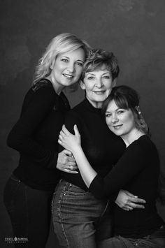 three women hugging each other while posing for a black and white photo in front of a dark background