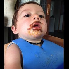 a young boy with food on his face in a blue bib, looking up at the camera