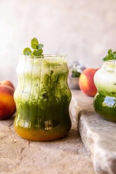 two glasses filled with green liquid sitting on top of a stone slab next to peaches