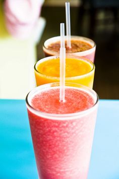 three glasses filled with different colored drinks on top of a blue table next to each other