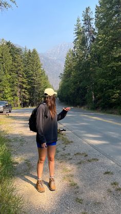 a woman standing on the side of a road pointing at something in the distance with trees and mountains behind her