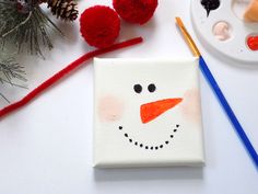 a snowman painted on a piece of canvas next to some paintbrushes and pine cones