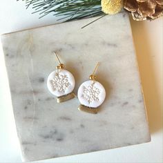 two white and gold earrings sitting on top of a marble slab next to pine cones