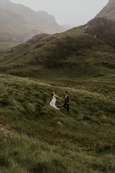 a bride and groom are walking through the grass on their wedding day in the mountains