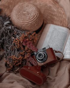 an open book, camera and hat laying on a bed next to a brown bag