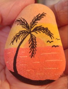 a hand holding a painted rock with a palm tree on the beach in the background