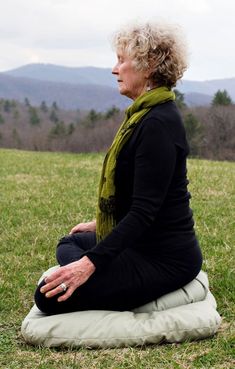 an older woman sitting on top of a pillow in the grass with mountains behind her