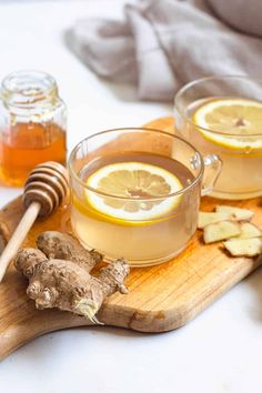 two mugs of tea with lemon and ginger on a cutting board next to honey