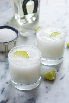 two glasses filled with white liquid and limes on a table next to salt and pepper shakers