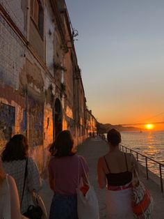 three women walking down a sidewalk next to the ocean at sunset or sunrise with an orange sun in the background