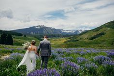 a bride and groom walking through the wildflowers with mountains in the back ground