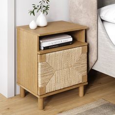 a wooden nightstand with books on it next to a white vase and bed in the background