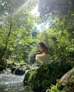 a woman sitting on top of a rock next to a stream in the forest with trees