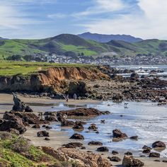 two people walking on the beach near some rocks and water with mountains in the background