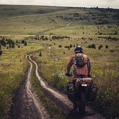 a man riding on the back of a motorcycle down a dirt road next to a lush green field