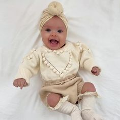 a baby is laying on a white sheet and wearing a knitted outfit with pom poms