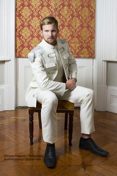 a man sitting on top of a wooden chair wearing a white suit and black shoes