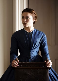 a woman in a blue dress is holding a wooden box and looking off into the distance