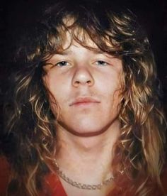 a man with long hair wearing a red shirt and chain around his neck is staring at the camera