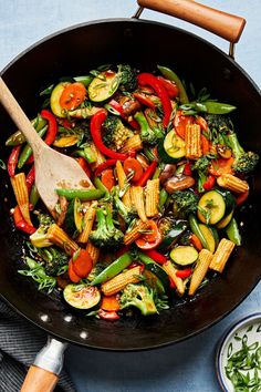 a skillet filled with stir fry vegetables on top of a blue table cloth next to a wooden spoon