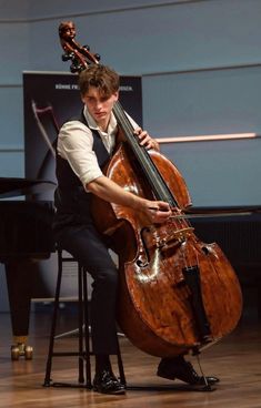 a young man sitting on a stool holding a large bass in his hands and playing the cello