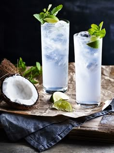 two glasses filled with ice and limes on top of a wooden table next to a coconut