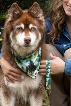 a woman is holding a dog wearing a bandana while sitting on the ground in front of trees