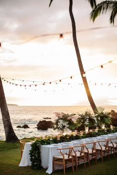 a long table set up with white linens and greenery on the beach at sunset