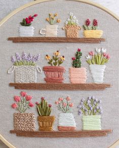 a cross stitched wall hanging with potted plants and flowers on it's shelves