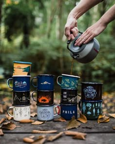 a person pours coffee from a teapot into several mugs in the woods