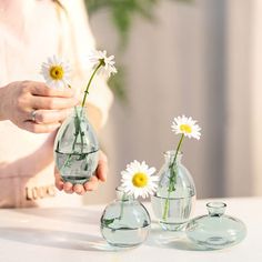 a woman is holding three vases with daisies in them on a white table