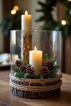 two candles are sitting in a glass container with pine cones and greenery on the table