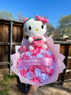 a hello kitty stuffed animal holding a bouquet of pink and purple roses in front of a wooden fence