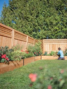 a woman sitting on the grass in front of a wooden fence and flower garden area