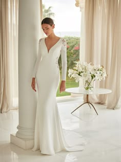 a woman standing in front of a window wearing a long sleeved wedding dress with an open back