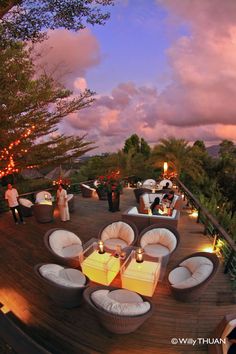 an outdoor dining area with white chairs and lights on the deck at dusk, surrounded by greenery