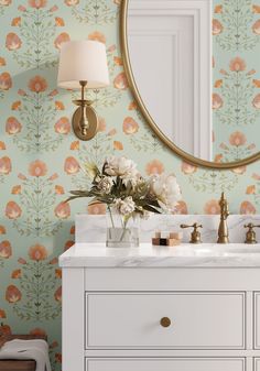 a bathroom vanity with flowers on the wall and a round mirror above it, along with a gold faucet