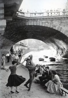 black and white photograph of people sitting on the side of a river under a bridge