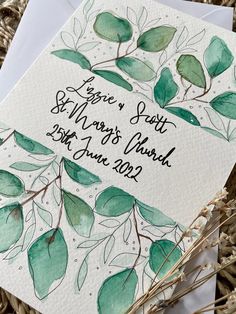 a wedding card with leaves on it and the date written in black ink, sitting next to some dried grass