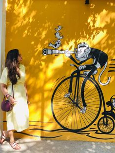 a woman standing next to a yellow wall with a painting of a monkey riding a bicycle