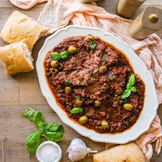 a white bowl filled with meat and olives on top of a wooden table next to bread