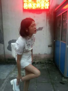 a woman squatting on the ground in front of a building with a neon sign above her