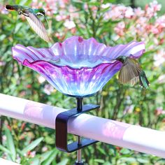 two hummingbirds flying over a purple flower bowl on a rail with pink flowers in the background