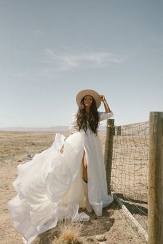 a woman in a white dress and hat leaning against a fence on the side of a dirt road